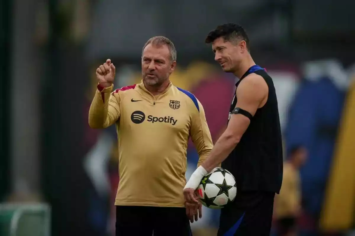 Dos hombres conversan en un campo de entrenamiento de fútbol, uno de ellos sostiene un balón y el otro señala algo mientras lleva una sudadera del FC Barcelona.