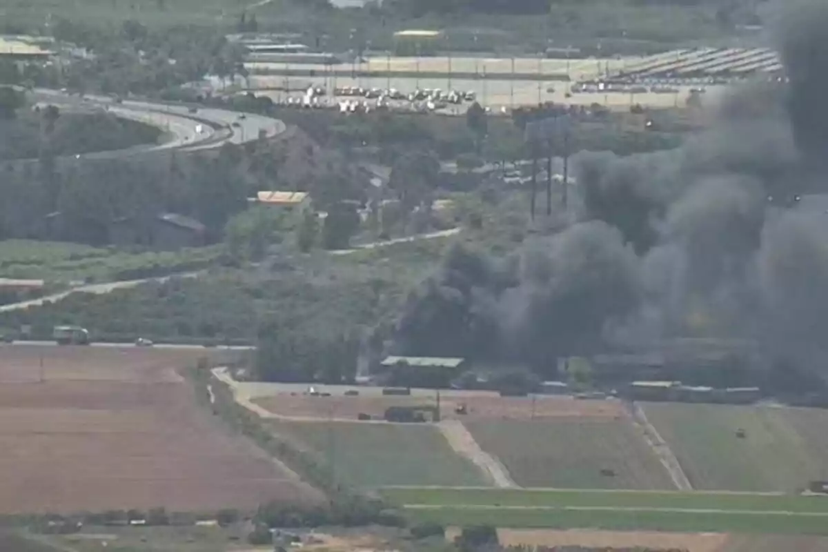 Núvol dens de fum negre s’aixeca des d’una zona industrial envoltada de camps i camins rurals. Núvol dens de fum negre s’aixeca des d’una zona industrial envoltada de camps i camins rurals.