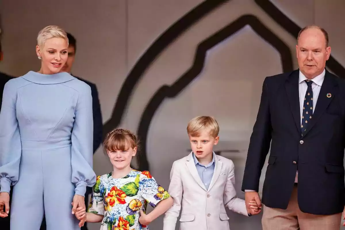 Una familia de cuatro personas, dos adultos y dos niños, posan juntos mientras se toman de las manos; la mujer lleva un vestido azul claro, la niña un vestido colorido con estampado floral, el niño un traje claro y el hombre un traje oscuro con corbata. Una familia de cuatro personas, dos adultos y dos niños, posan juntos mientras se toman de las manos; la mujer lleva un vestido azul claro, la niña un vestido colorido con estampado floral, el niño un traje claro y el hombre un traje oscuro con corbata.