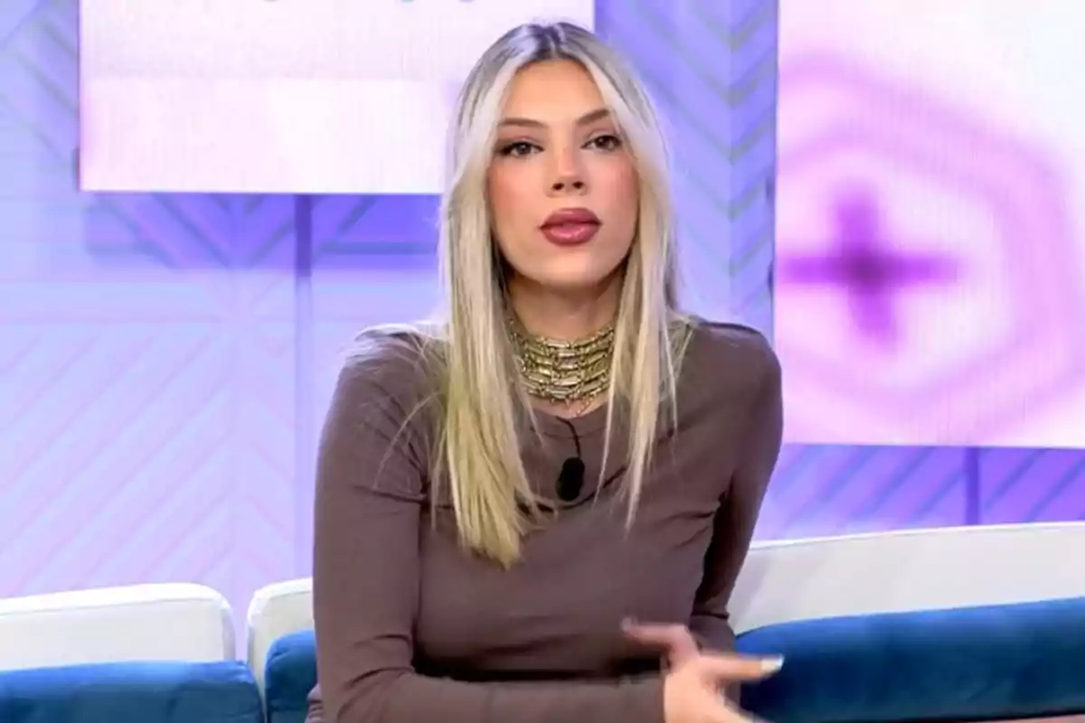 Alejandra Rubio con cabello rubio y largo está sentada en un sofá durante un programa de televisión, lleva un vestido marrón y un collar dorado, el fondo es de tonos morados y azules. Alejandra Rubio con cabello rubio y largo está sentada en un sofá durante un programa de televisión, lleva un vestido marrón y un collar dorado, el fondo es de tonos morados y azules.