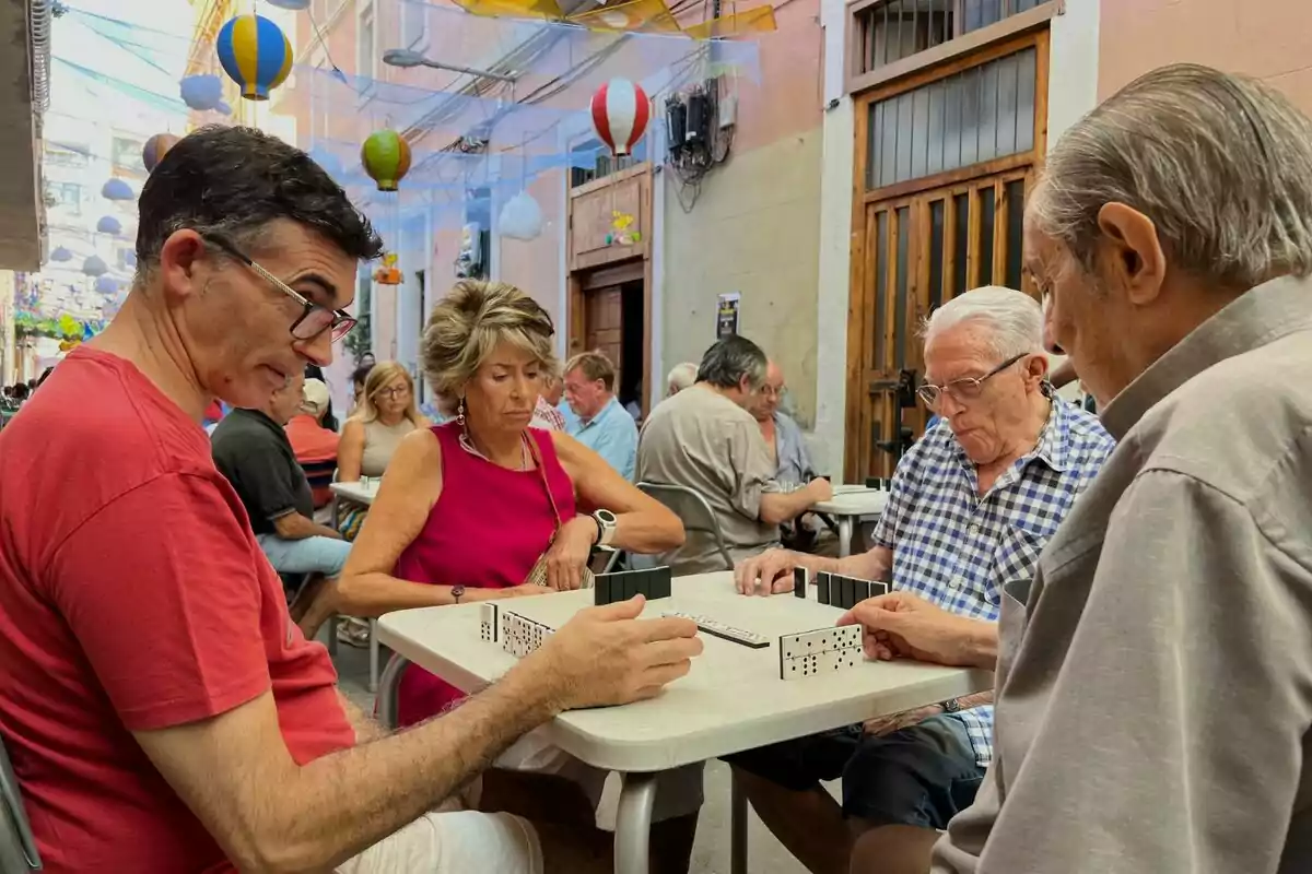 Un grup de persones juga al dòmino en una terrassa exterior decorada amb globus aerostàtics de colors.