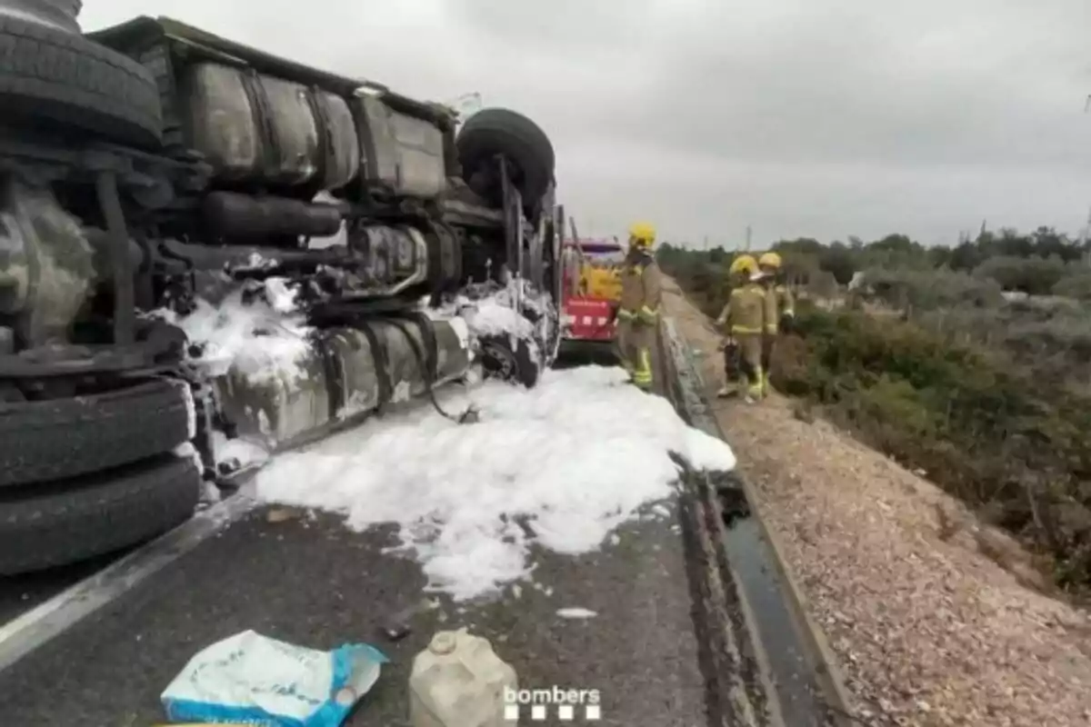 Un camió bolcat a la carretera amb escuma al voltant mentre bombers treballen al lloc.