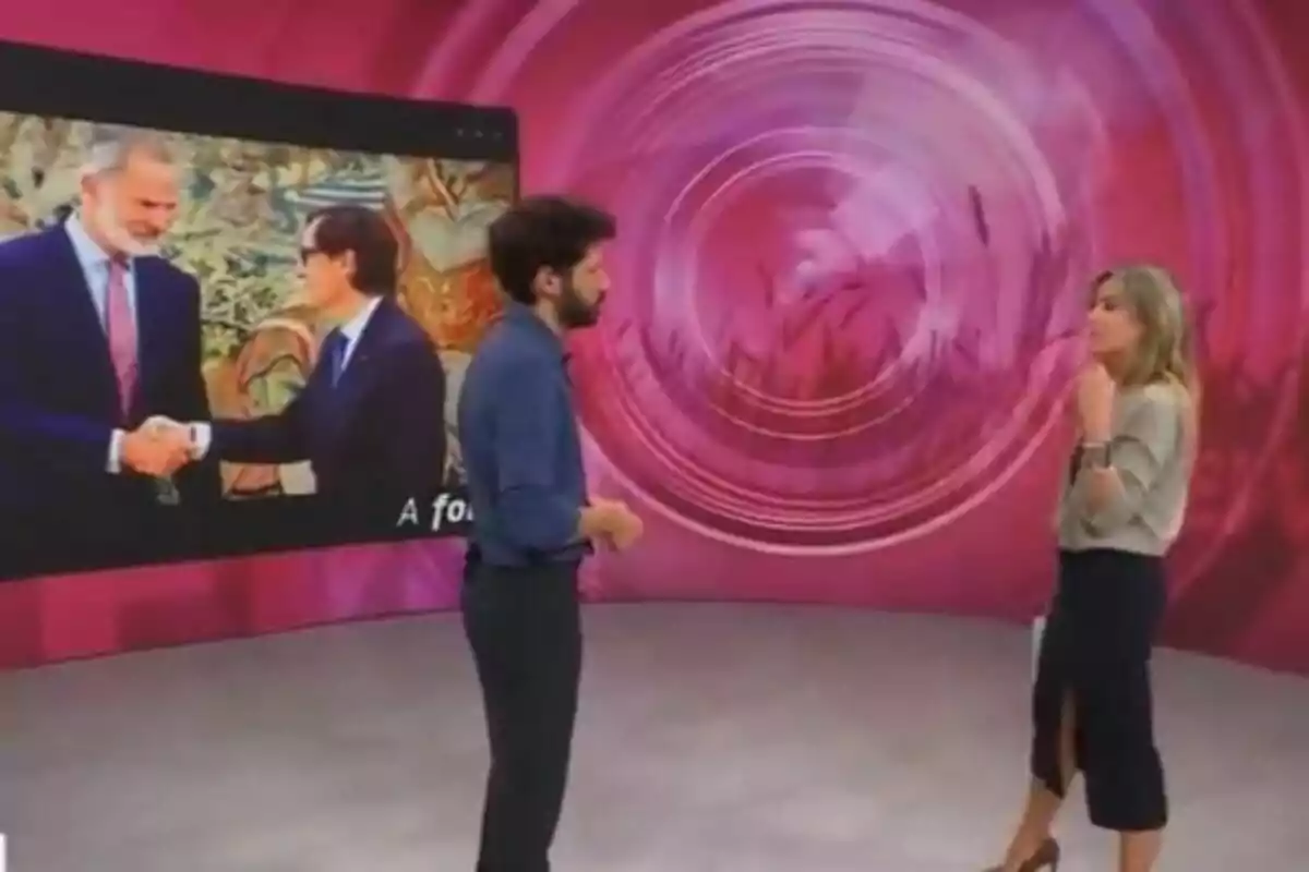 Dos personas conversan en un estudio de televisión con un fondo rosa y una pantalla que muestra a dos hombres dándose la mano.