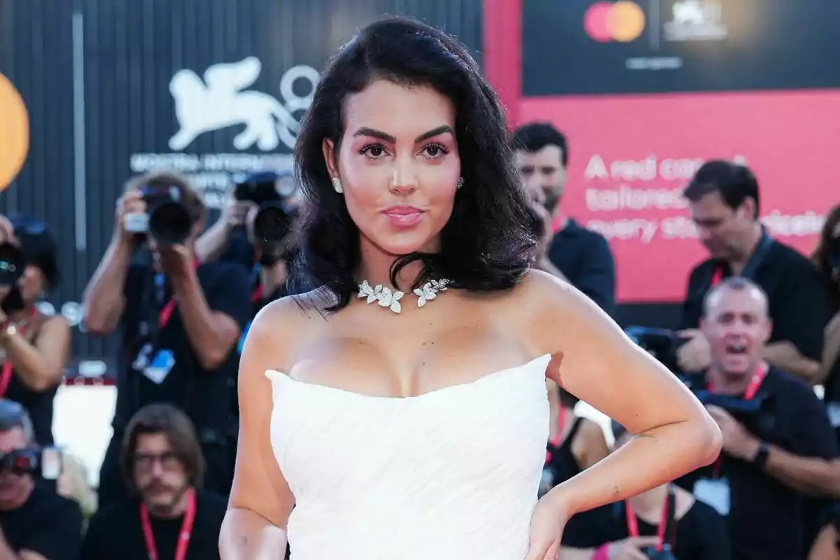 Dona amb vestit blanc elegant i collaret de flors posant en una catifa vermella mentre fotògrafs la retraten al fons Dona amb vestit blanc elegant i collaret de flors posant en una catifa vermella mentre fotògrafs la retraten al fons
