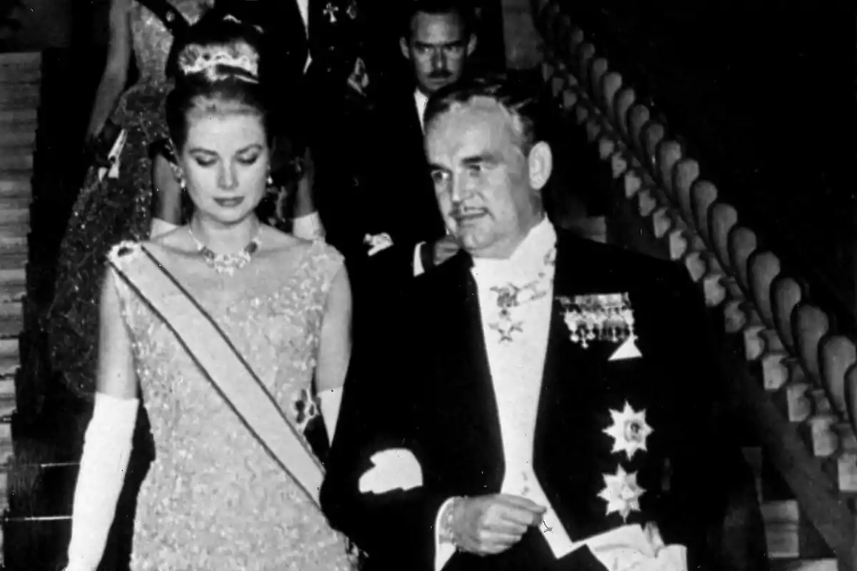 Una mujer con vestido elegante y tiara camina junto a un hombre con traje formal y condecoraciones en un evento de gala.