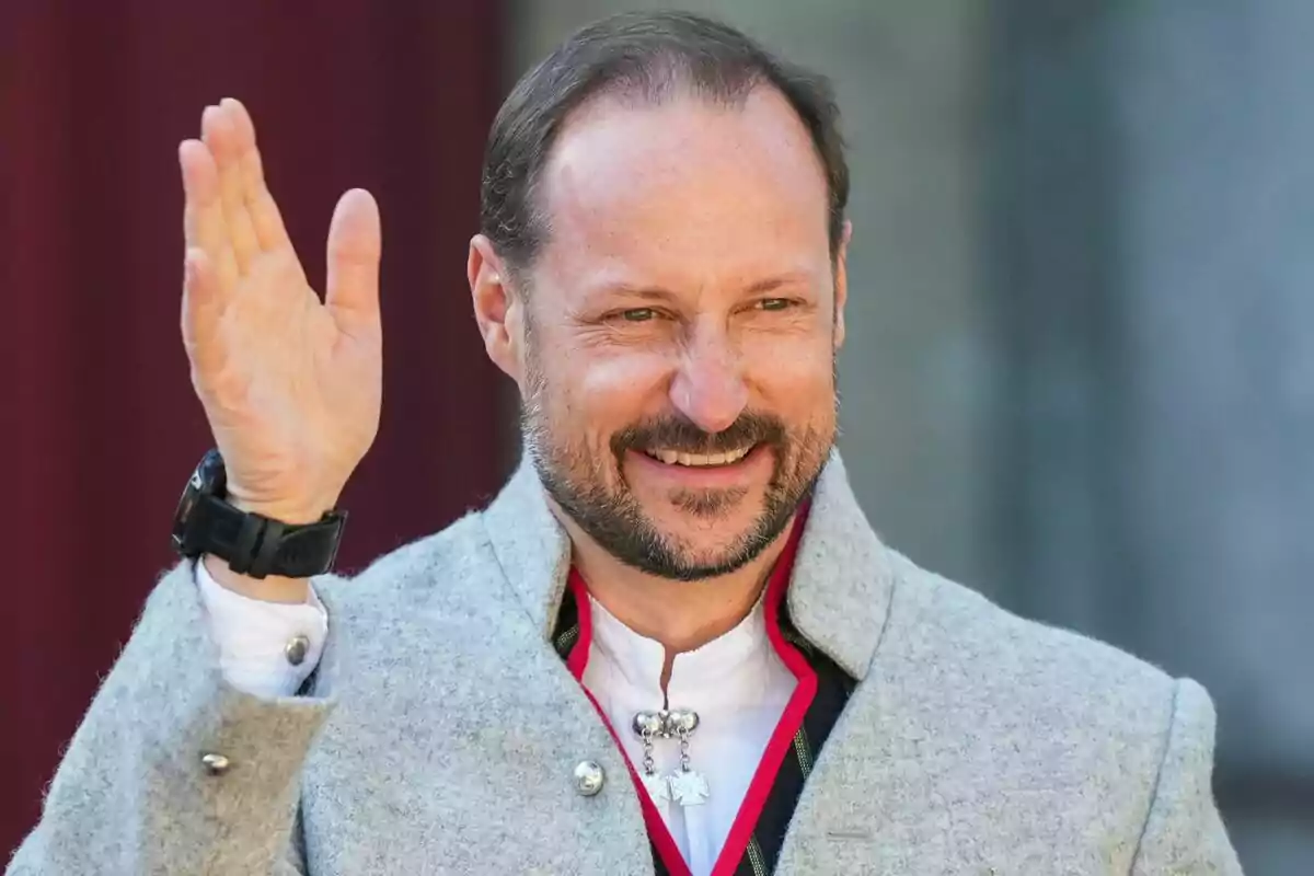 Un hombre sonriente con barba y bigote, vistiendo un abrigo gris y saludando con la mano levantada. Un hombre sonriente con barba y bigote, vistiendo un abrigo gris y saludando con la mano levantada.