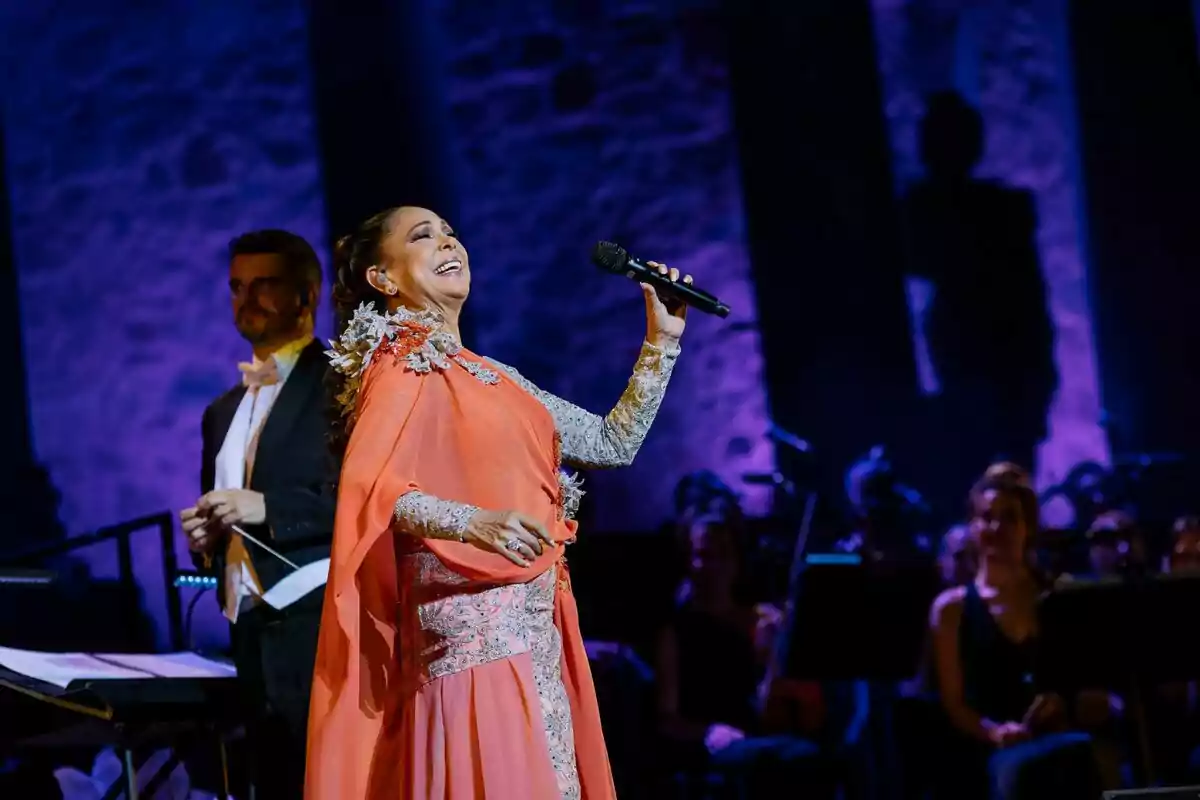 Una dona amb un vestit elegant canta en un escenari il·luminat de color porpra mentre sosté un micròfon. Una dona amb un vestit elegant canta en un escenari il·luminat de color porpra mentre sosté un micròfon.