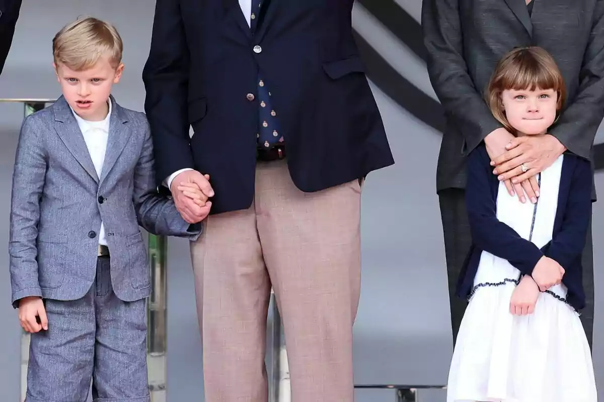 Un niño y una niña están de pie junto a dos adultos, el niño sostiene la mano de uno de los adultos mientras la niña está abrazada por el otro adulto. Un niño y una niña están de pie junto a dos adultos, el niño sostiene la mano de uno de los adultos mientras la niña está abrazada por el otro adulto.