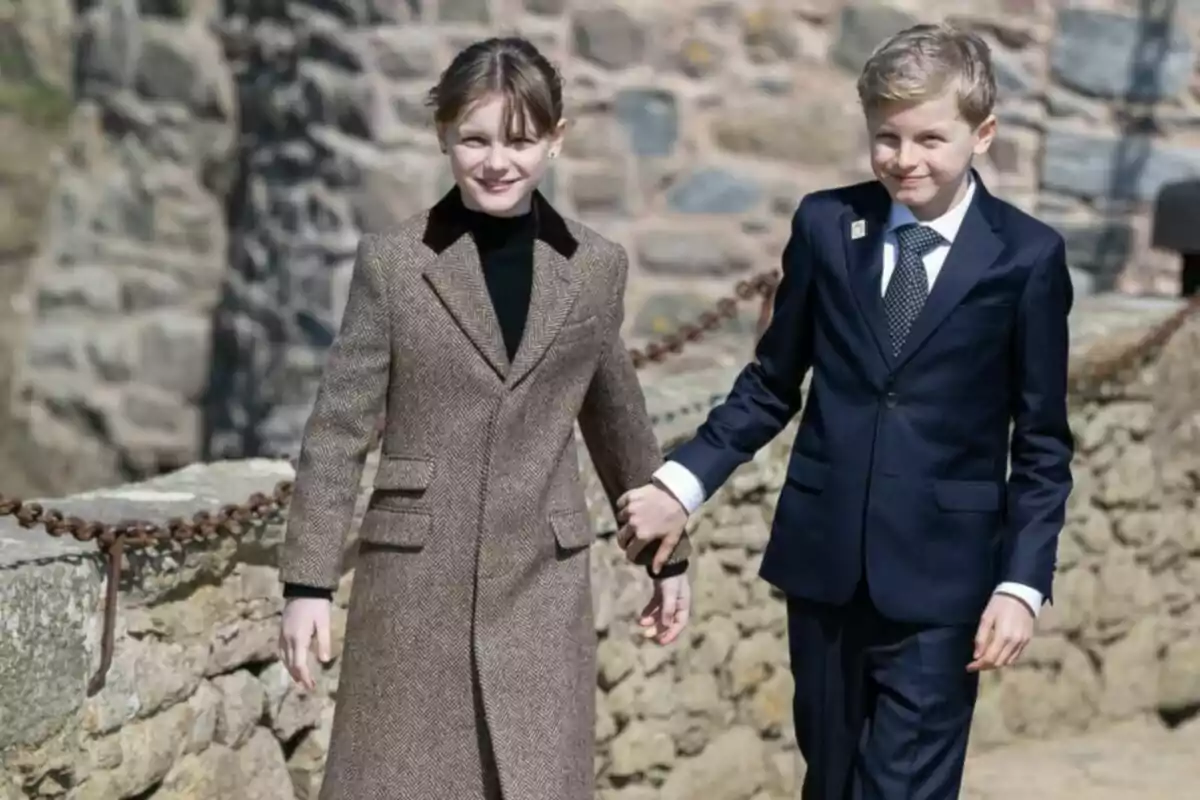 Two well-dressed children walking hand in hand in an outdoor setting with a stone wall in the background. Two well-dressed children walking hand in hand in an outdoor setting with a stone wall in the background.