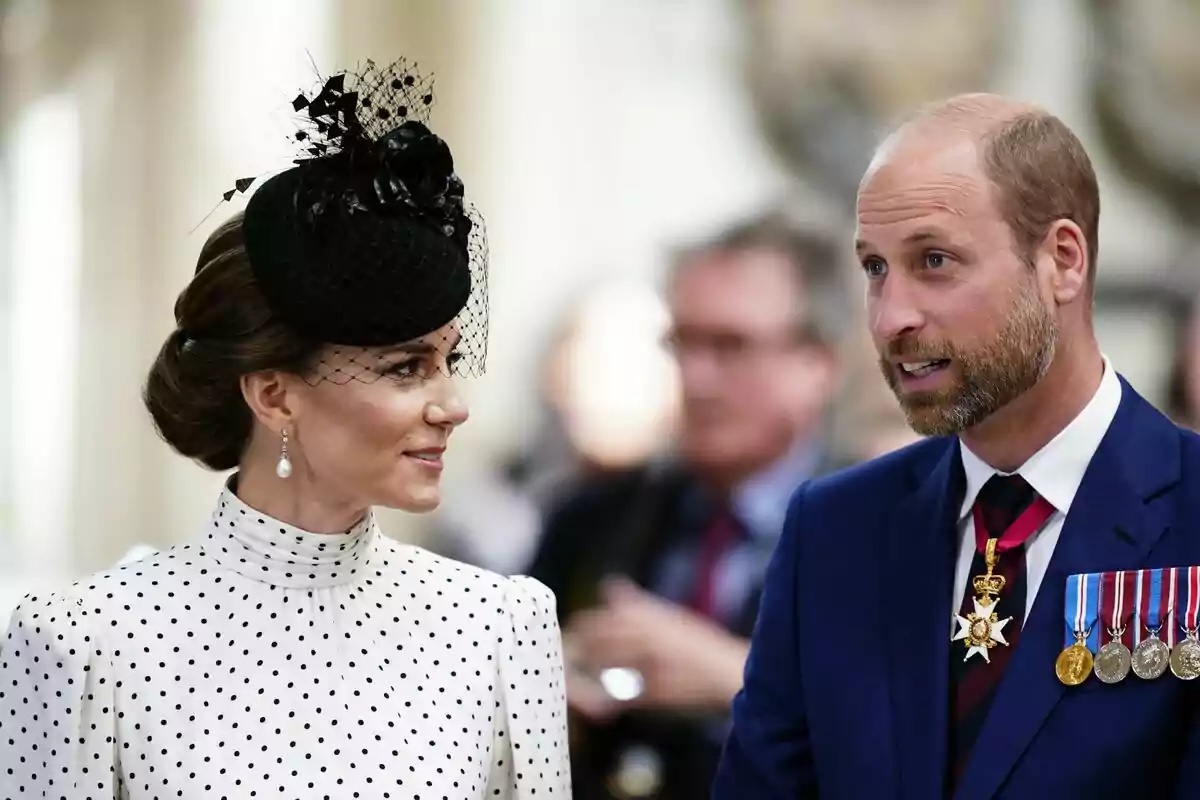 Una mujer con un elegante sombrero negro y vestido de lunares conversa con un hombre que lleva un traje azul y varias medallas.