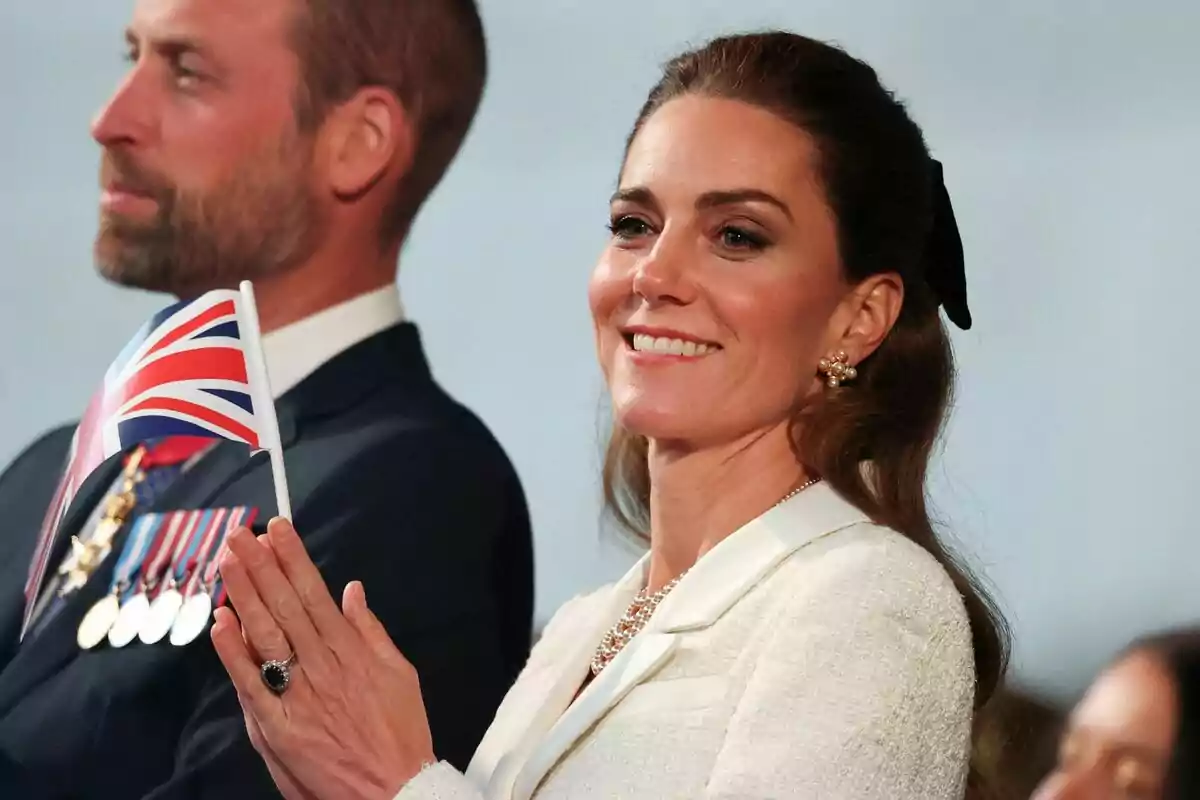 Una dona somrient sosté una petita bandera del Regne Unit mentre està dreta al costat d'un home amb medalles al seu vestit. Una dona somrient sosté una petita bandera del Regne Unit mentre està dreta al costat d'un home amb medalles al seu vestit.