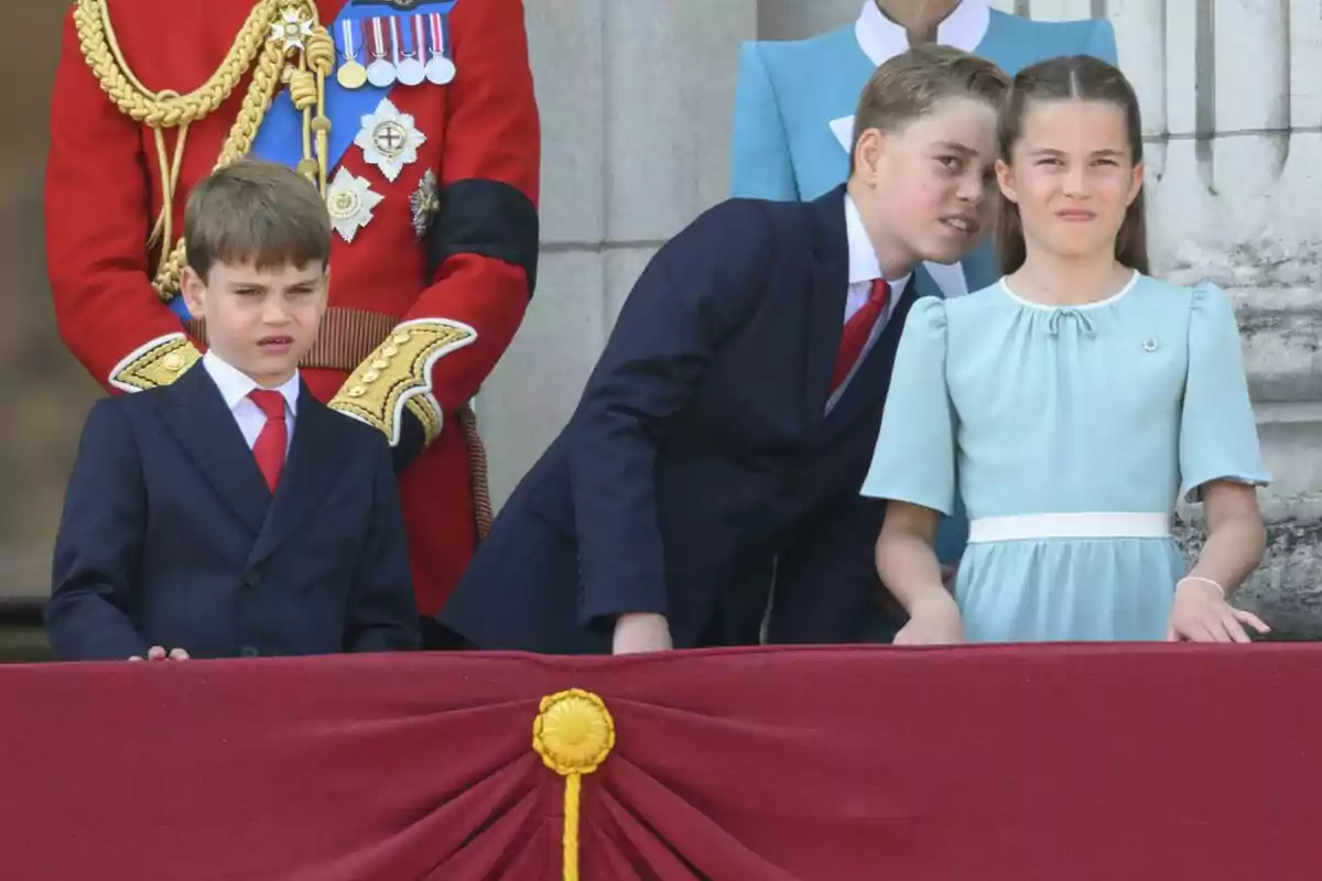 Tres niños vestidos de manera formal observan un evento desde un balcón decorado con tela roja y detalles dorados, mientras adultos elegantemente vestidos están detrás de ellos. Tres niños vestidos de manera formal observan un evento desde un balcón decorado con tela roja y detalles dorados, mientras adultos elegantemente vestidos están detrás de ellos.