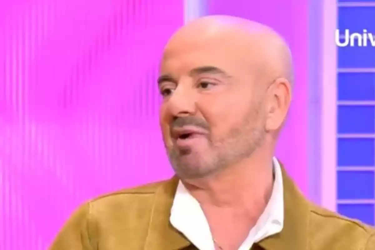Un home calb amb barba parlant en un programa de televisió amb fons rosa.