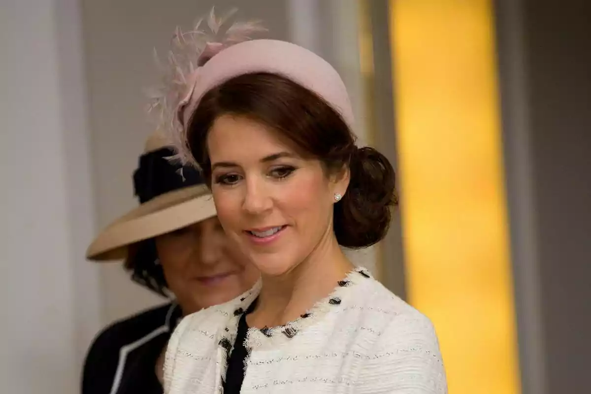 Una dona amb un elegant barret rosa i un pentinat recollit somriu mentre porta un abric clar amb detalls foscos.