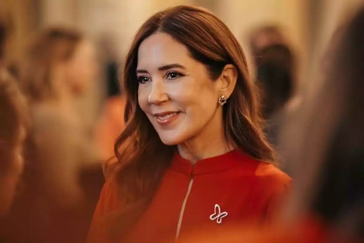 A woman with loose brown hair, wearing a red blouse with a butterfly-shaped brooch, smiles while surrounded by people at an event. A woman with loose brown hair, wearing a red blouse with a butterfly-shaped brooch, smiles while surrounded by people at an event.