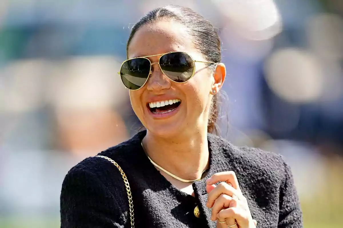 Mujer sonriente con gafas de sol y chaqueta oscura en un ambiente exterior luminoso Mujer sonriente con gafas de sol y chaqueta oscura en un ambiente exterior luminoso