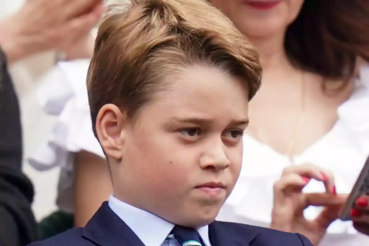 A boy with a serious expression, dressed in a suit and tie, surrounded by people at an event. A boy with a serious expression, dressed in a suit and tie, surrounded by people at an event.