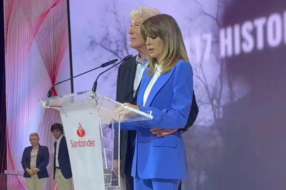 Dues persones dretes darrere d’un faristol amb el logotip de Santander, una d’elles llegint mentre l’altra l’acompanya, en un escenari amb fons il·luminat i dues persones més al fons. Dues persones dretes darrere d’un faristol amb el logotip de Santander, una d’elles llegint mentre l’altra l’acompanya, en un escenari amb fons il·luminat i dues persones més al fons.