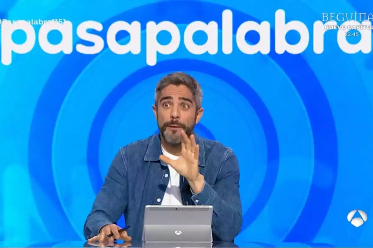 Un presentador amb barba i jaqueta de mezclilla està davant d'una pantalla blava amb el logotip de Un presentador amb barba i jaqueta de mezclilla està davant d'una pantalla blava amb el logotip de
