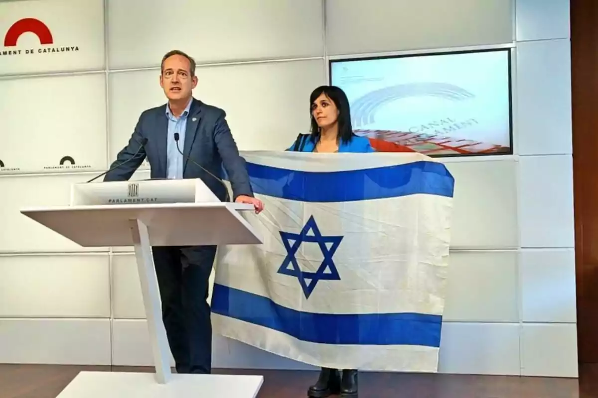 Un home amb vestit parla en un faristol amb el logotip del Parlament de Catalunya mentre una dona sosté una bandera d'Israel al seu costat.