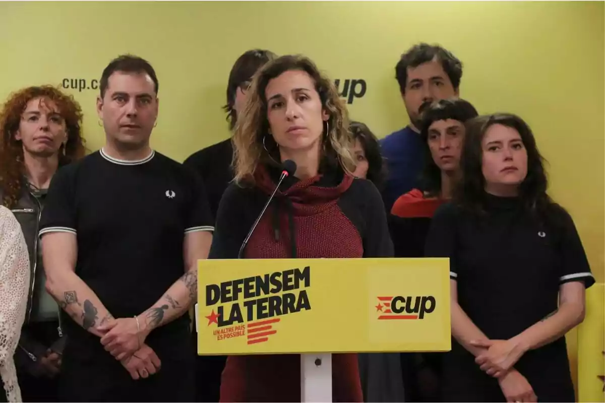 Un grup de persones està dret davant d'un fons groc amb un cartell que diu "Defensem la terra" i "cup", mentre una dona parla en un micròfon.