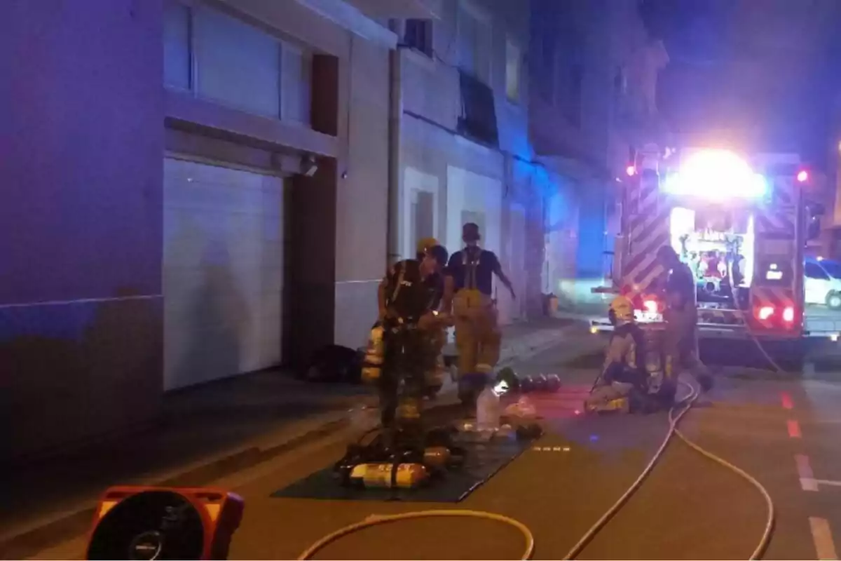 Bombers treballant en un carrer durant la nit amb equip de respiració i una unitat d’emergència il·luminada al fons