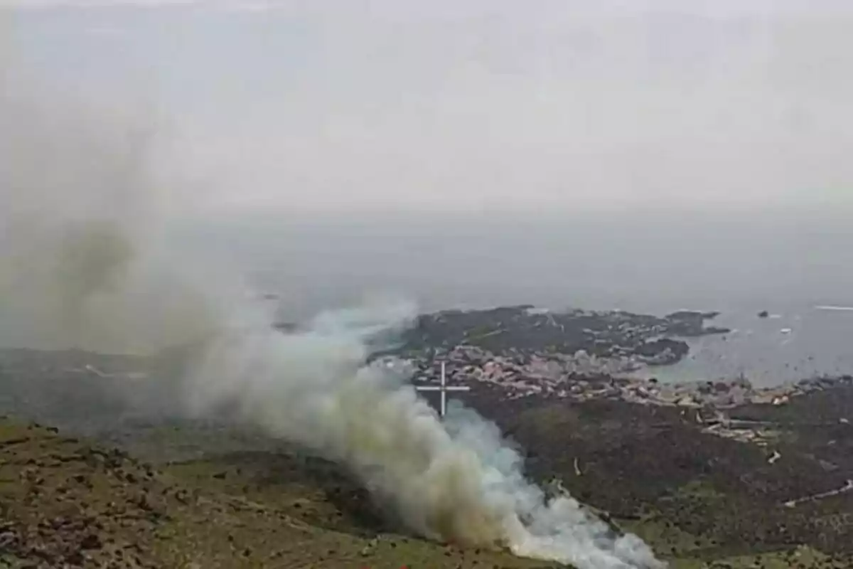 Fum dens s’eleva d’un incendi en una zona muntanyosa a prop de la costa i d’un poble vora el mar. Fum dens s’eleva d’un incendi en una zona muntanyosa a prop de la costa i d’un poble vora el mar.