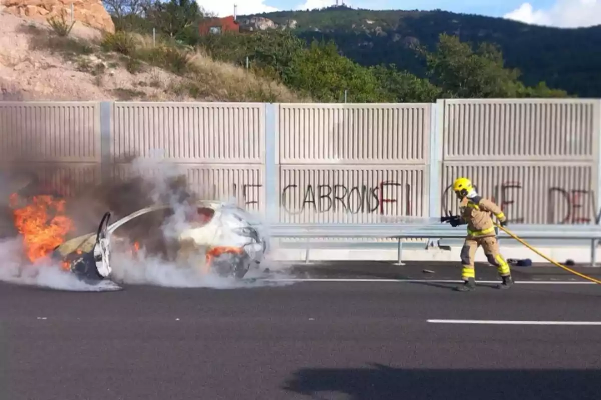 Un bombero se acerca a un coche en llamas en una carretera, con un muro de contención al fondo.