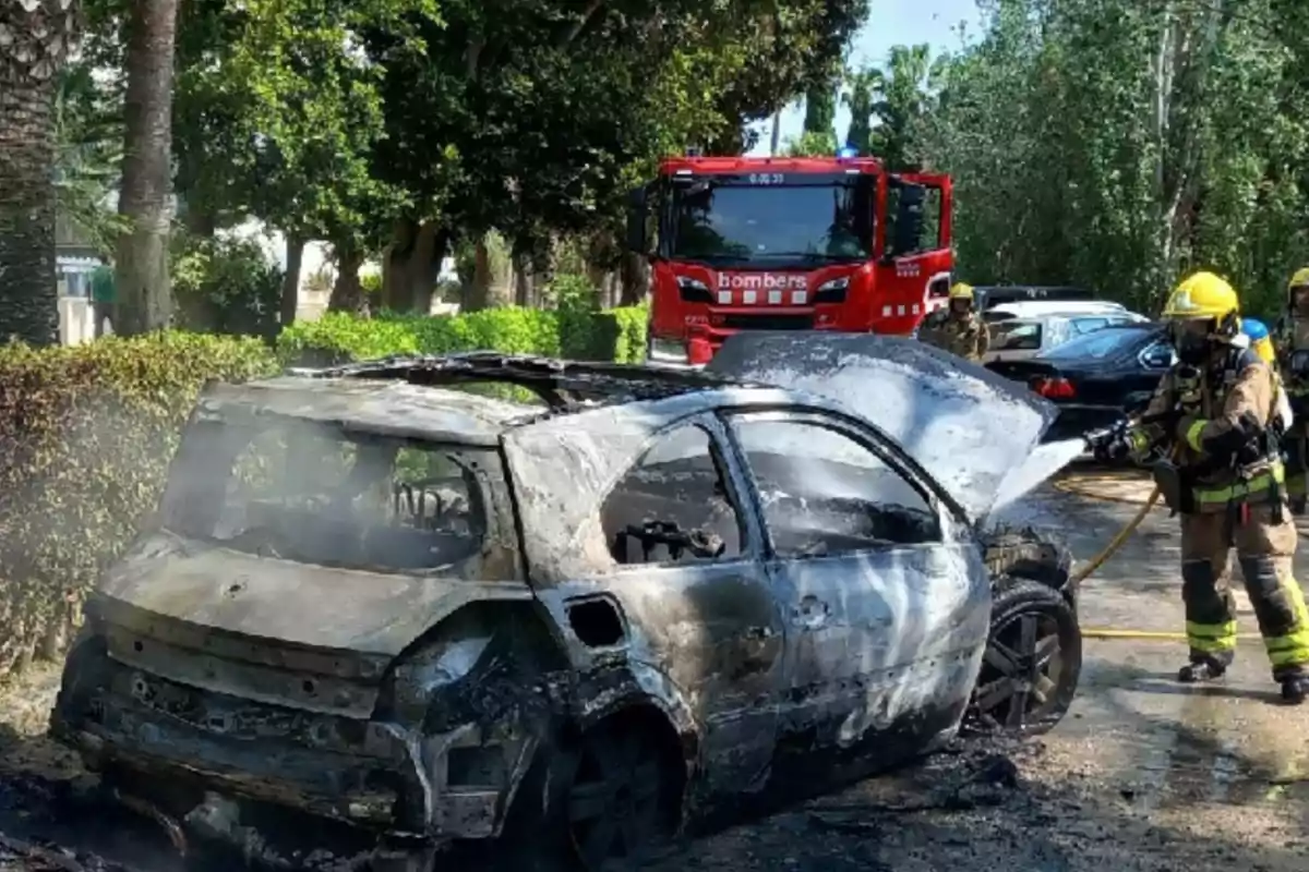 Un cotxe completament calcinat amb bombers treballant per apagar el foc i un camió de bombers al fons