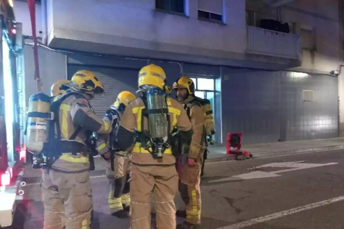 Bomberos con equipo de protección y tanques de oxígeno conversan frente a un edificio durante la noche.