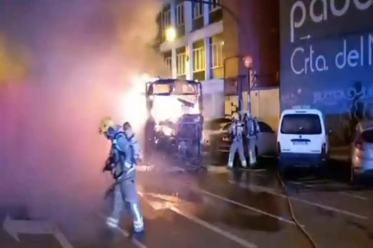 Bomberos combaten un incendio en un autobús en una calle urbana durante la noche con humo denso y luces de emergencia iluminando la escena.