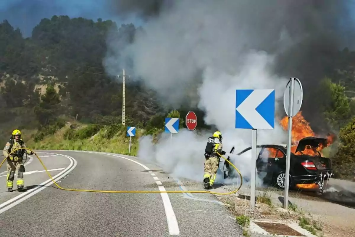 Dos bomberos apagan un incendio en un coche en llamas al costado de una carretera rodeada de vegetación y señales de tráfico