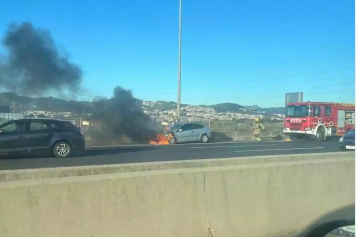 Un cotxe gris en flames en una carretera amb fum negre sortint del capó mentre un camió de bombers i un bomber són a prop.