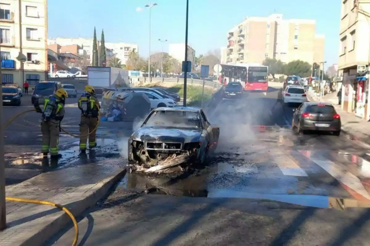 Bomberos apagando un coche incendiado en una calle urbana con varios vehículos y edificios alrededor.