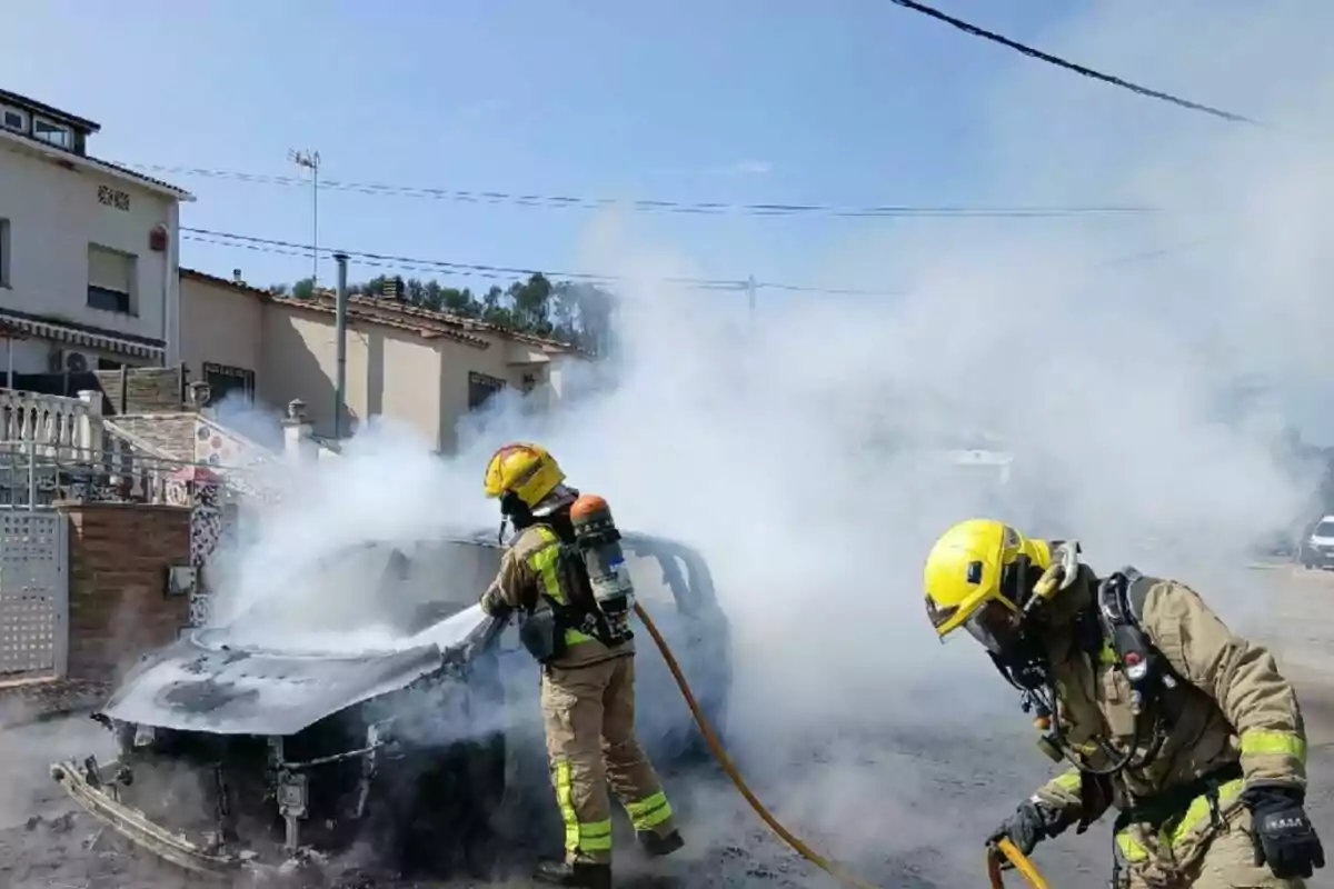 Bombers apagant un cotxe en flames en una zona residencial, amb fum dens al voltant.