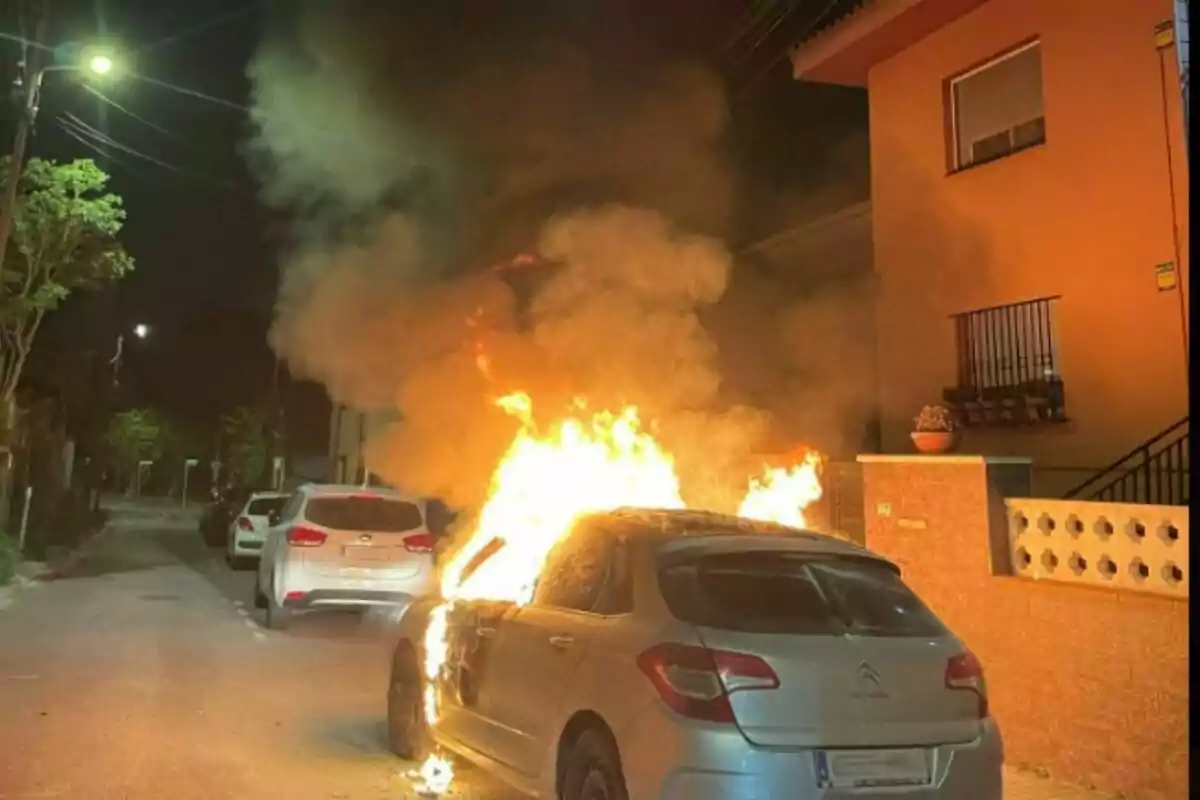 Un cotxe en flames en un carrer residencial durant la nit, amb fum dens elevant-se cap al cel.