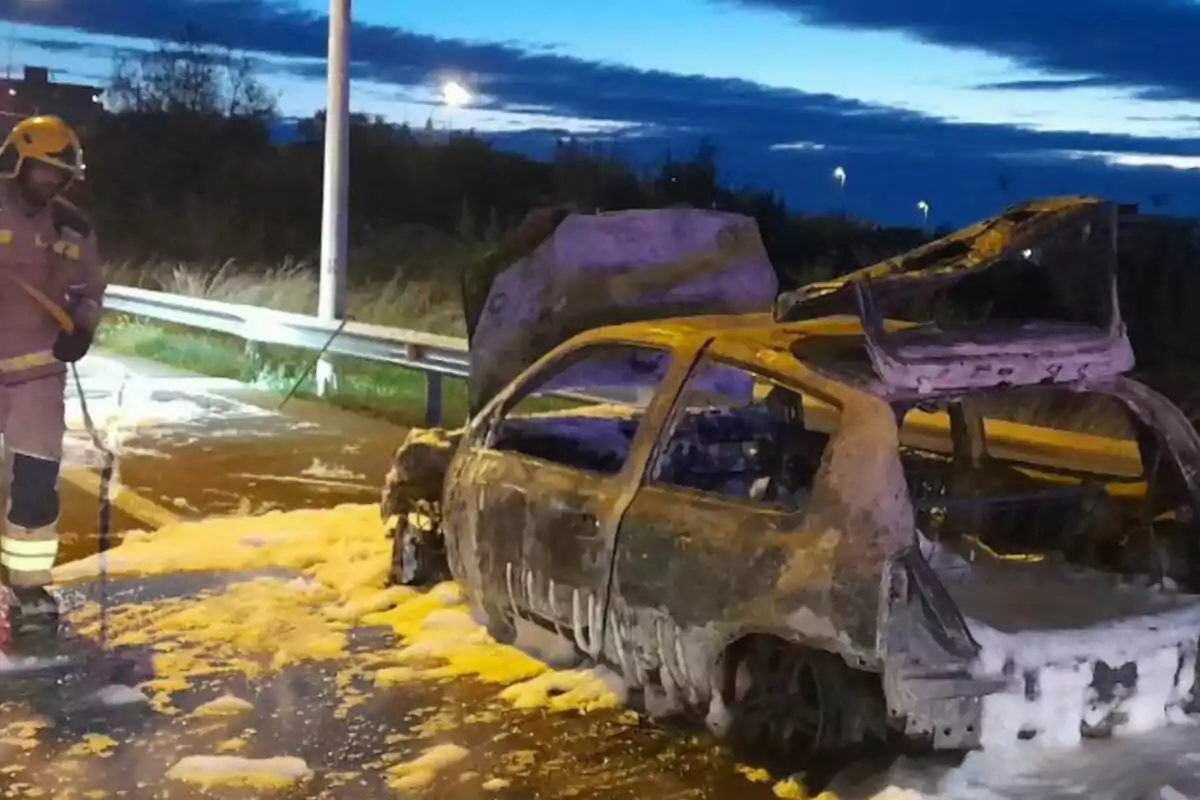 Un bomber al costat d'un cotxe cremat cobert d'escuma en una carretera al capvespre.