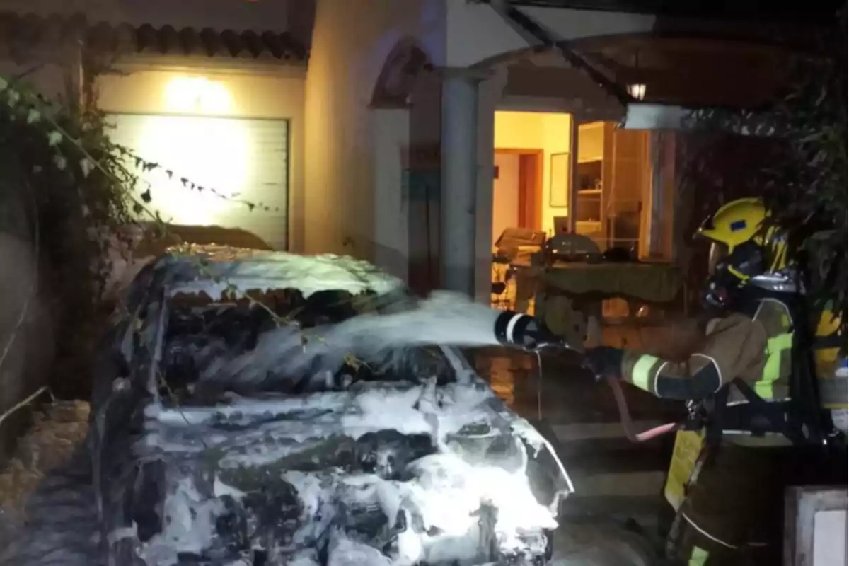 Un bombero apaga un incendio en un automóvil con espuma frente a una casa iluminada.