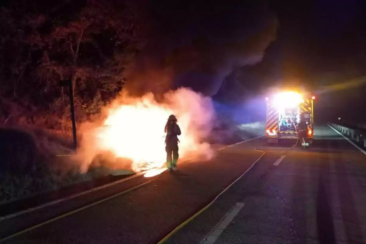 Bomber treballant per apagar un incendi a la carretera durant la nit amb un camió de bombers il·luminat al fons. Bomber treballant per apagar un incendi a la carretera durant la nit amb un camió de bombers il·luminat al fons.