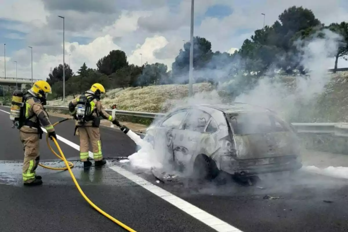 Dos bomberos con equipo completo apagan un incendio en un automóvil en una carretera usando espuma mientras el vehículo emite humo.