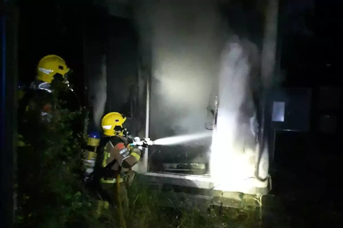 Bombers amb casc groc apaguen un incendi en un edifici durant la nit fent servir una mànega d’aigua.