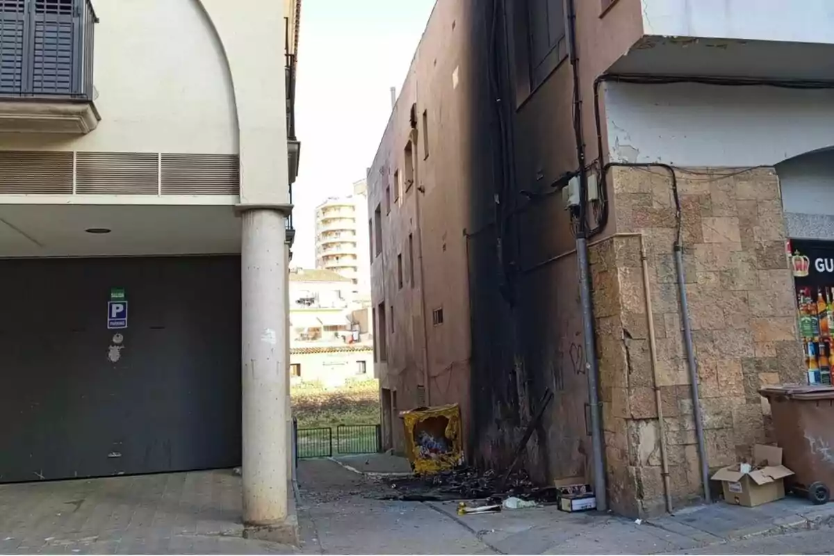 Contenidor d’escombraries cremat al costat d’un edifici amb la paret enfosquida pel fum i restes d’escombraries escampades per terra.