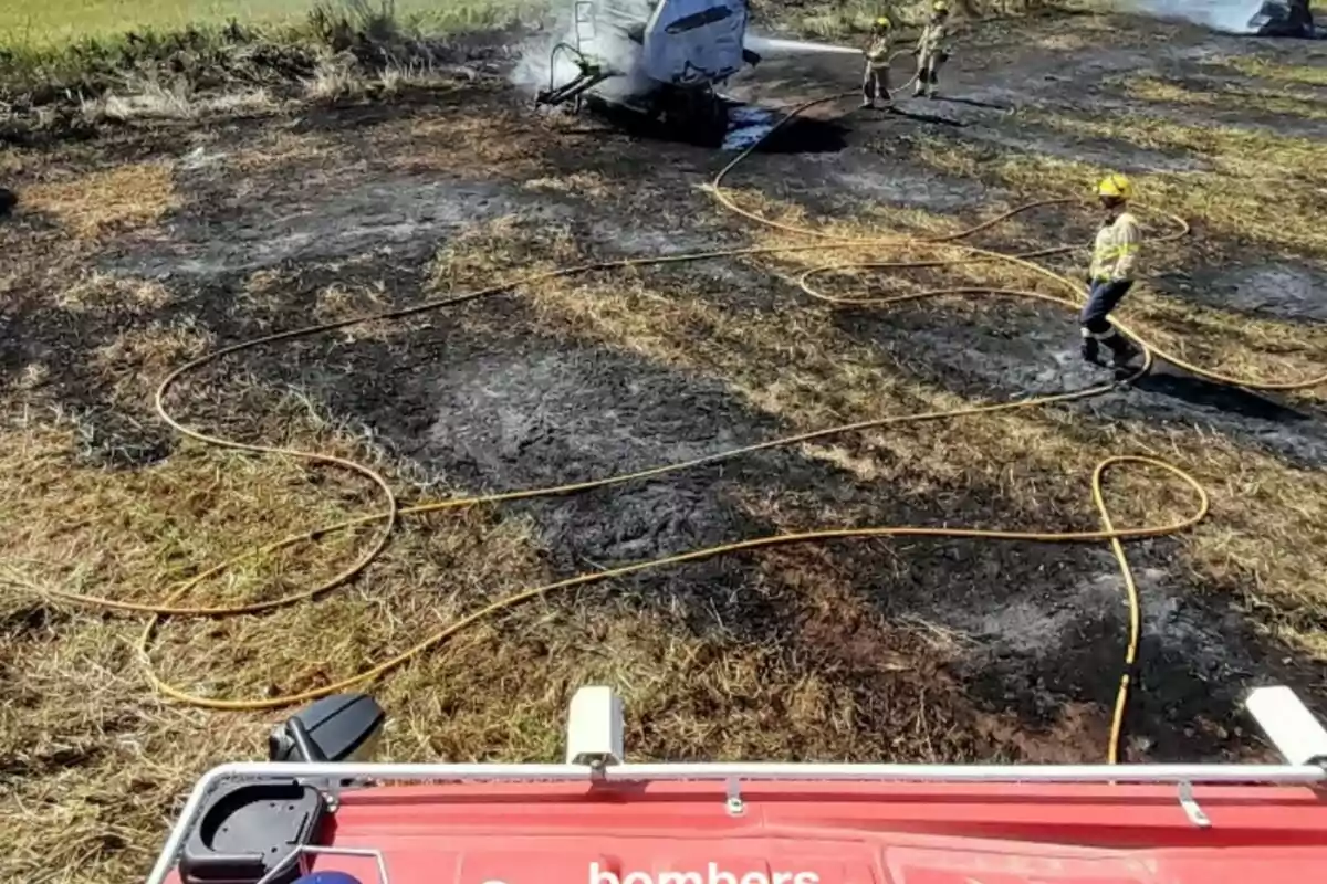 Bombers treballant en un camp cremat amb mànegues esteses i un vehicle d'emergència en primer pla.