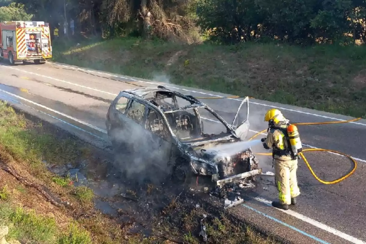Un bomber apaga l’incendi d’un cotxe completament calcinat en una carretera mentre un camió de bombers està estacionat al fons.