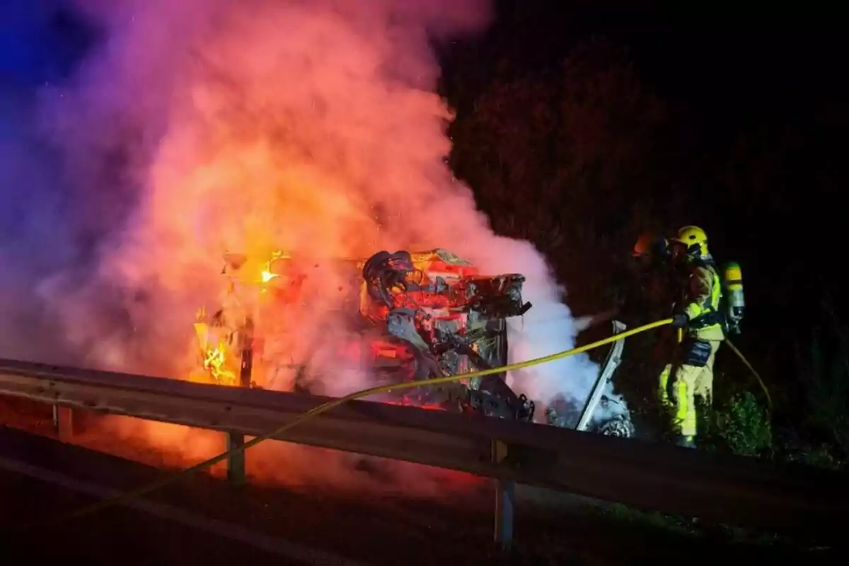 Bomberos extinguiendo un incendio en un vehículo volcado al lado de una carretera por la noche.