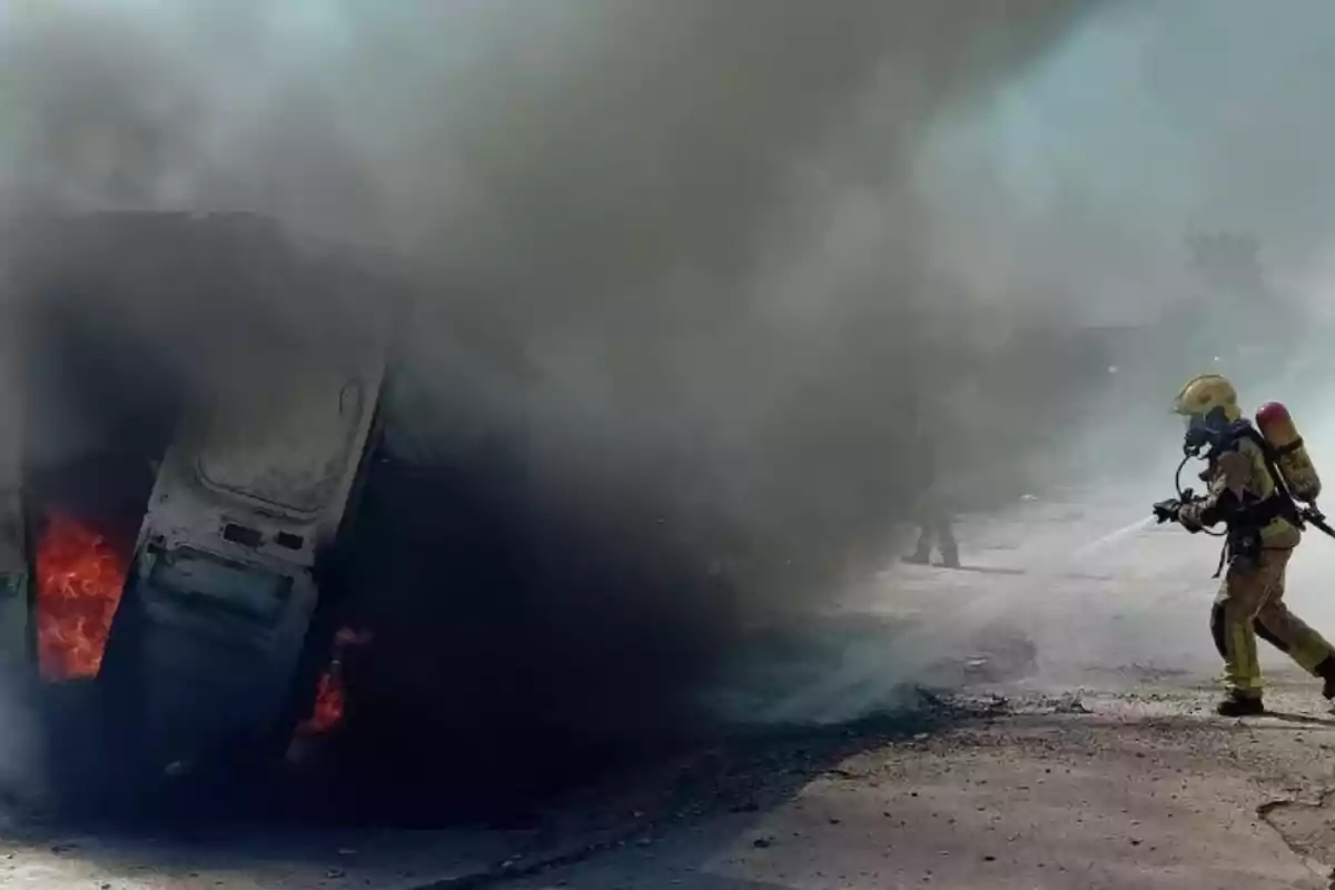 Un bomber apaga l’incendi d’un vehicle bolcat mentre un núvol dens de fum cobreix l’escena.