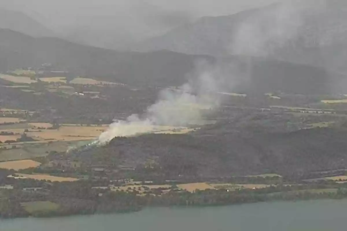 Fum blanc s’aixeca des d’una zona boscosa en un paisatge rural amb muntanyes al fons i un llac en primer terme.