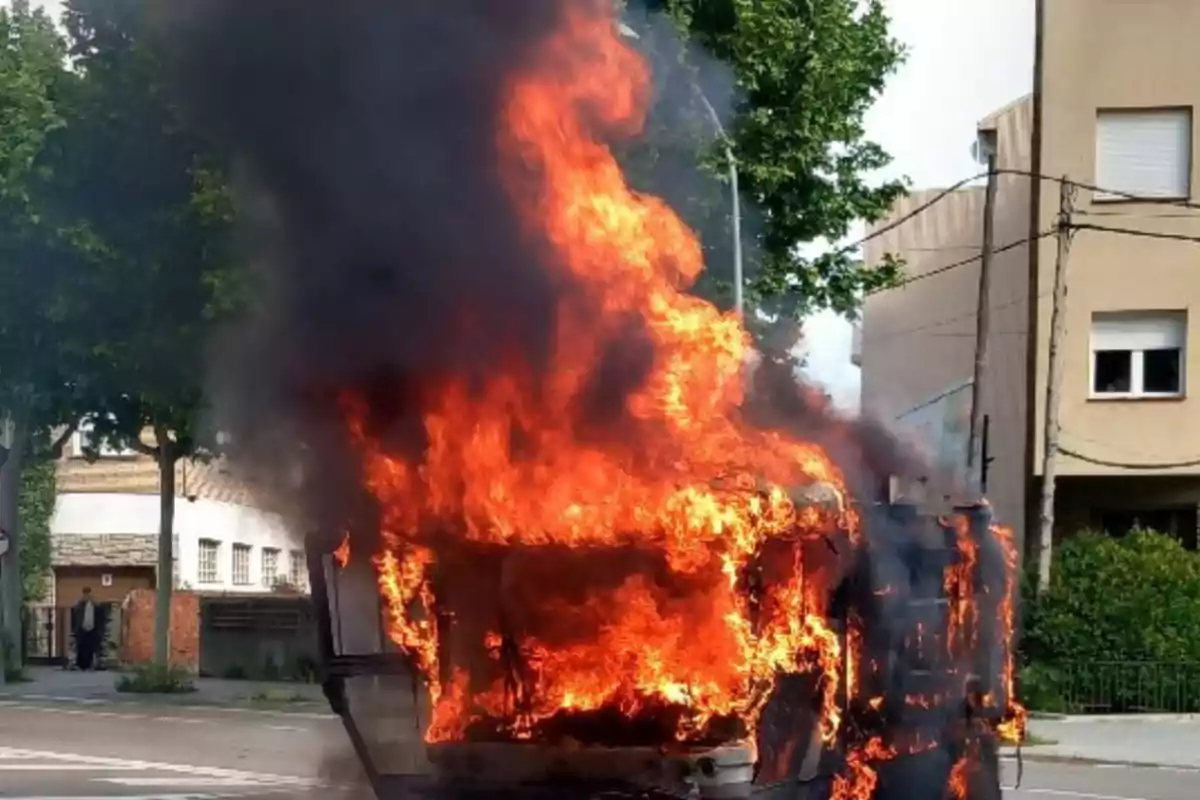 Un vehicle en flames en un carrer urbà amb arbres i edificis de fons.