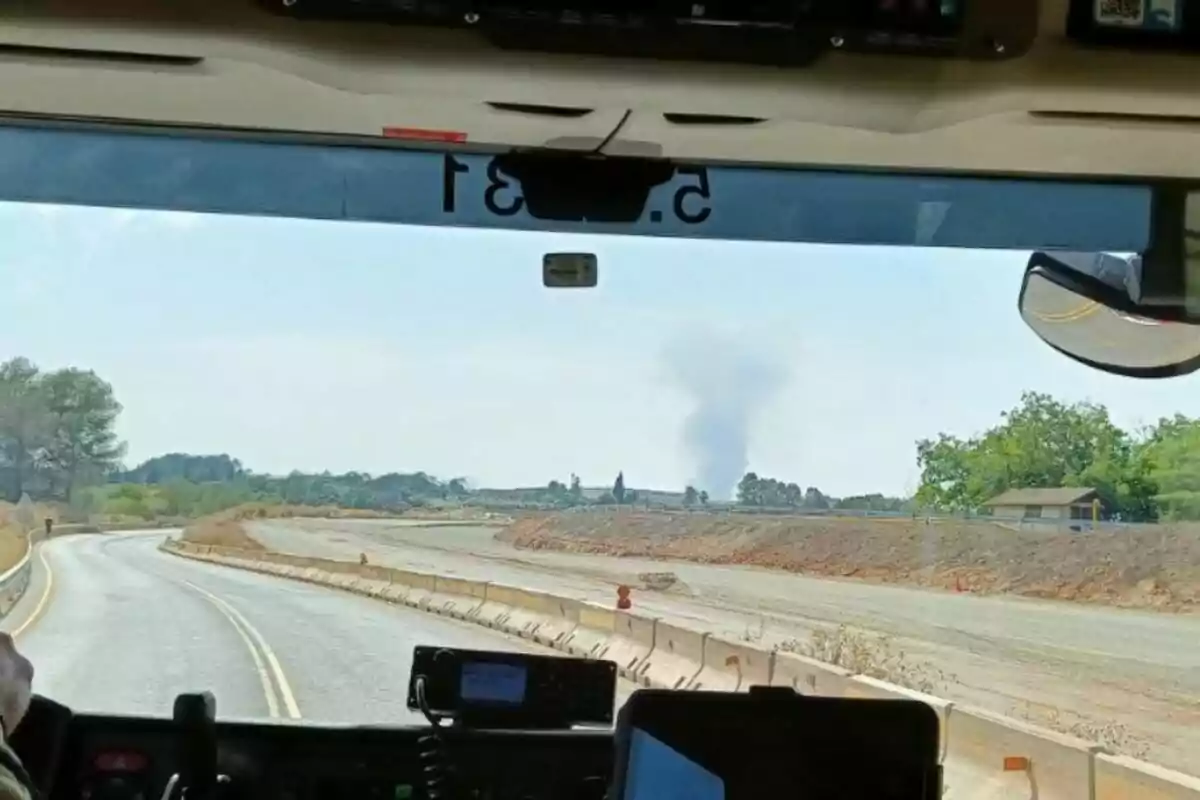 Vista des de l'interior d'un vehicle que circula per una carretera en construcció amb una columna de fum visible a la llunyania a l'horitzó