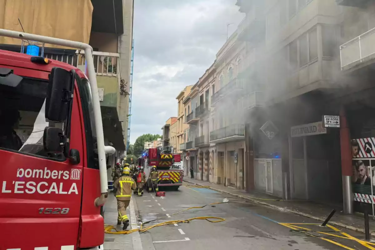 Camió de bombers i diversos bombers treballant en un carrer d’on surt fum d’un edifici.